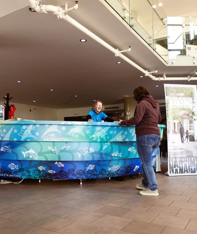 A visitor speaks with staff at a tourism information desk inside the Nanaimo Museum, with promotional displays nearby.