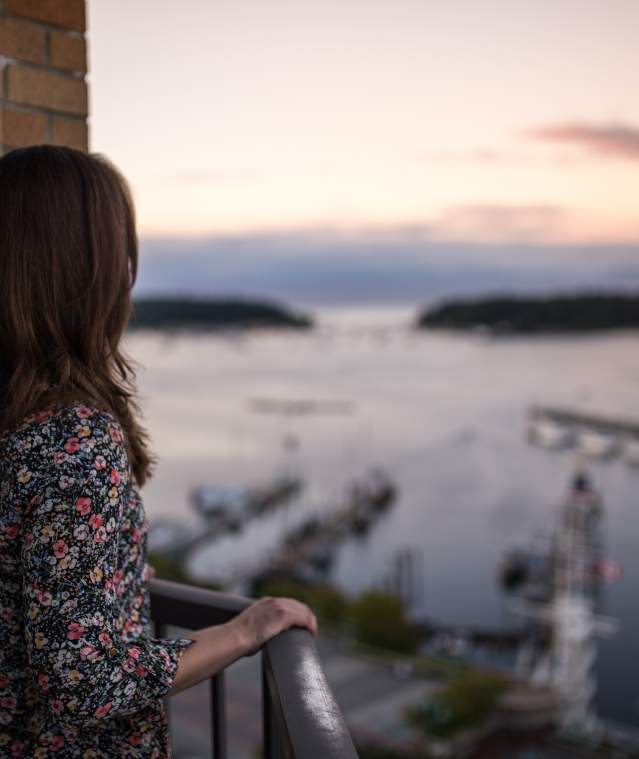 A person stands on a balcony overlooking a harbour with boats and distant islands at sunset.