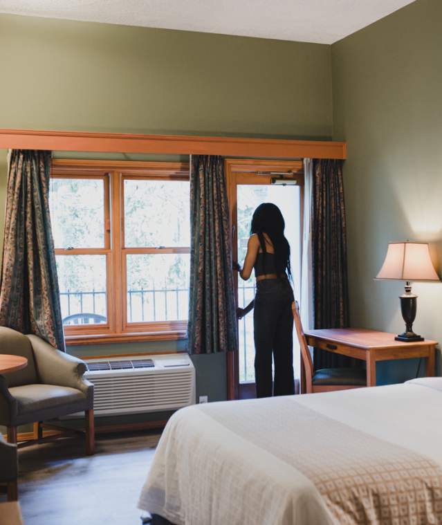 A cozy hotel room with a bed, desk, and seating area as a person opens a balcony door to let in natural light.
