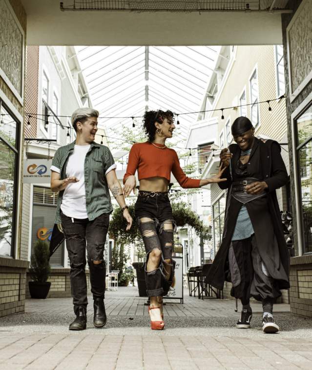 Three friends walk side by side, laughing in a bright, covered shopping walkway lined with storefronts.