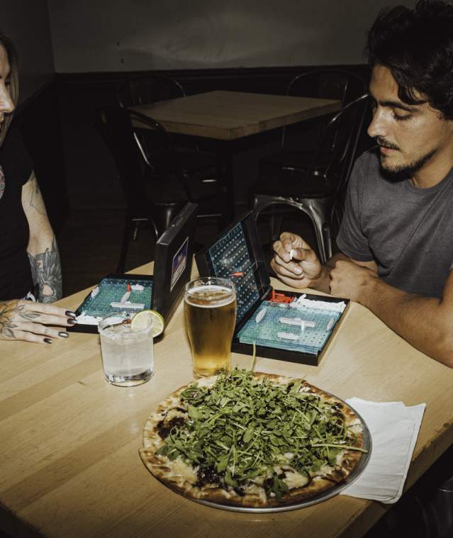 Two people play a tabletop game at a restaurant table with pizza and drinks in front of them.
