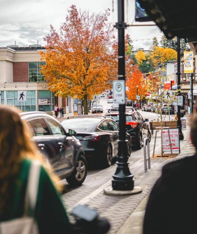 People walk along a downtown Nanaimo street with shops, parked cars, and vibrant autumn trees lining the road.