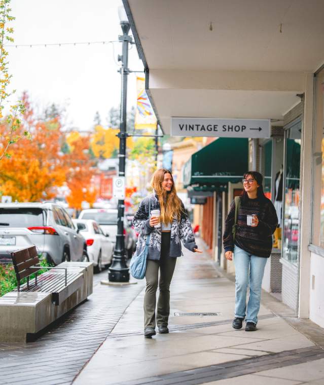Two people stroll past shops on a rainy autumn day in downtown Nanaimo, holding coffee as colourful fall trees line the street.