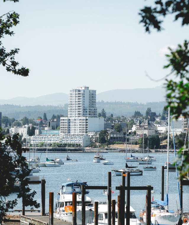 A view of Nanaimo’s waterfront with anchored boats and marina in the foreground, city skyline and distant hills visible through tree branches.
