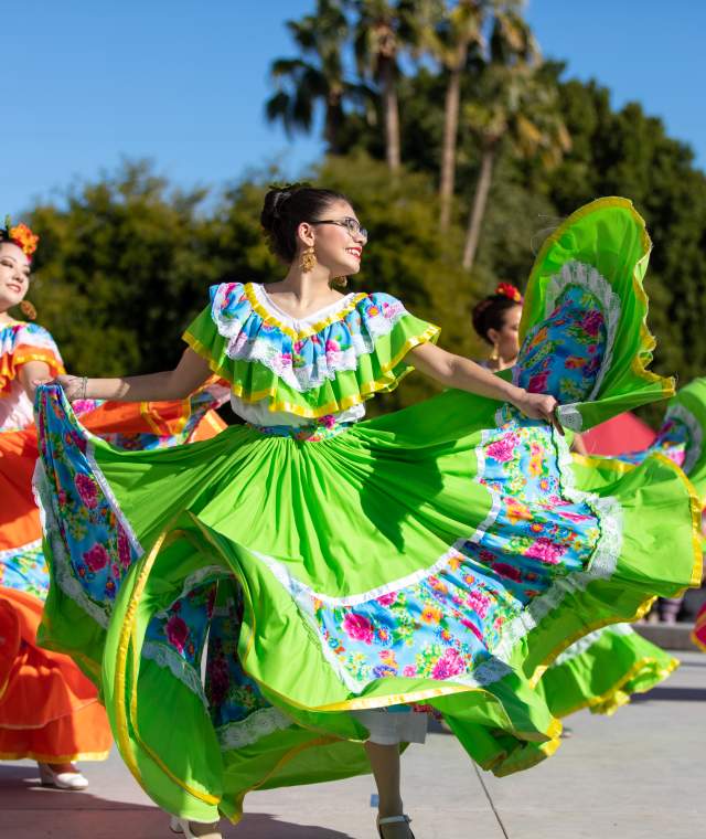 three dancers wearing brightly colored, floral dresses performing a traditional Folkloric dance honoring their Mexican history at an outdoor event