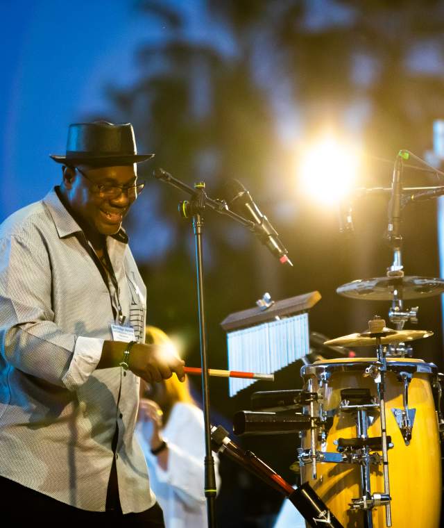 smiling musician playing instruments on an outdoor stage with trees in the background