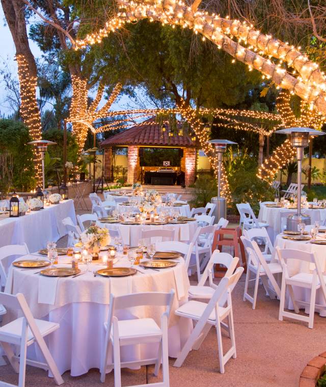 Elegant outdoor wedding setup with lit palm trees, white tables adorned with floral centerpieces, and a cozy ambiance.