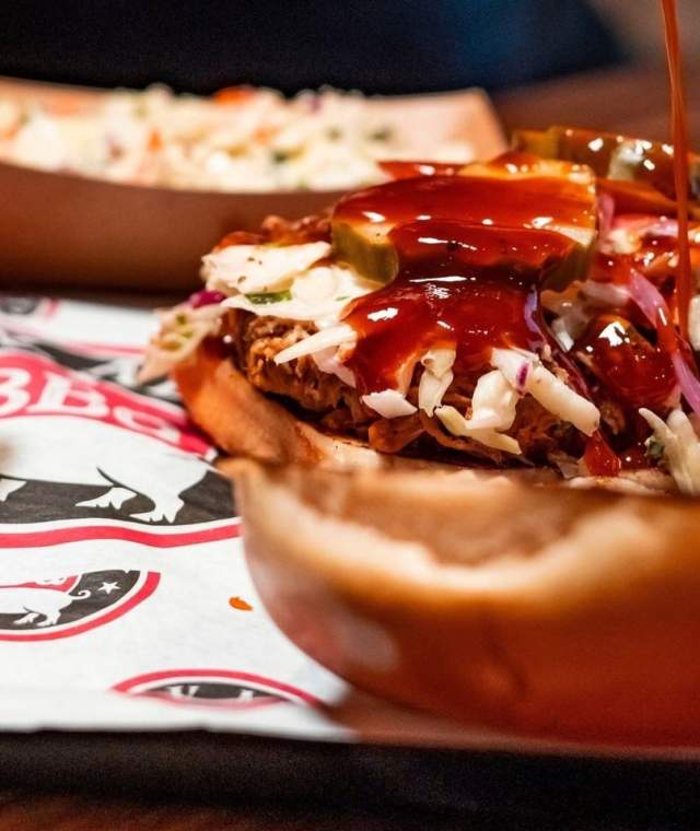 BBQ sandwich with bun open and BBQ sauce being drizzled on it. Sides are blurred in the background and food is sitting on a tray lined with Back Alley BBQ branded paper.