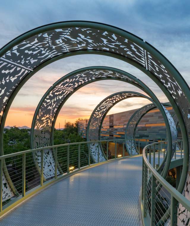 The Pecos Bridge, an elevated walkway at Chandler Gilbert Community College, features an artistic, molded metal structure around the perimeter with lights at the base and a sunset in the background.