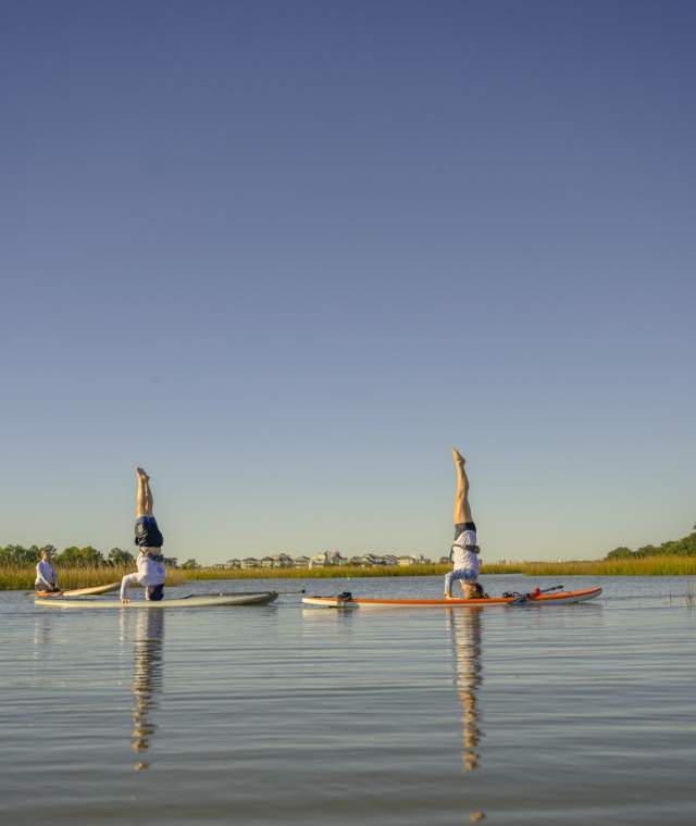 Women practicing paddleboard yoga with headstands on calm marsh waters in North Myrtle Beach, South Carolina, under a clear blue sky surrounded by peaceful natural scenery.