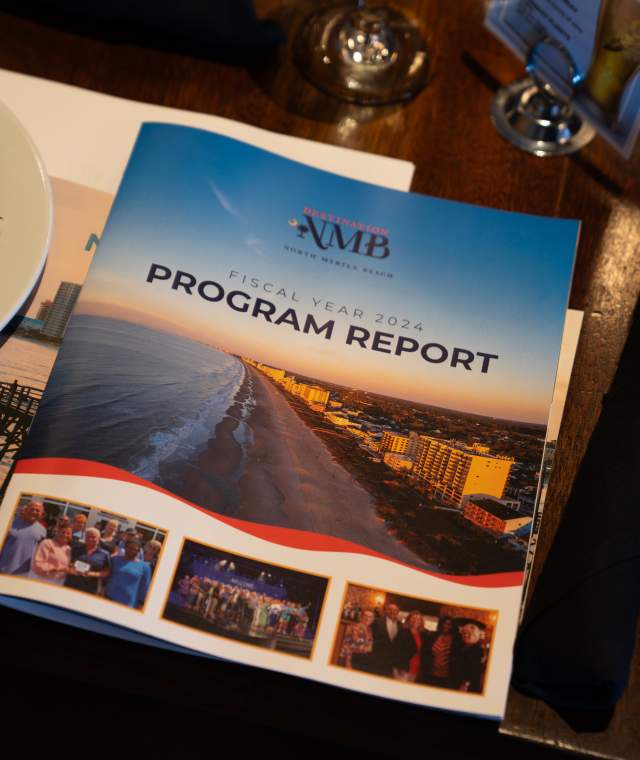 Destination NMB Program Report” booklet is displayed on a dark wooden dining table between two plates of salad. The cover features an aerial view of the North Myrtle Beach coastline and photos of community events. Wine glasses, silverware, and a black cloth napkin surround the report, suggesting a professional luncheon or banquet setting