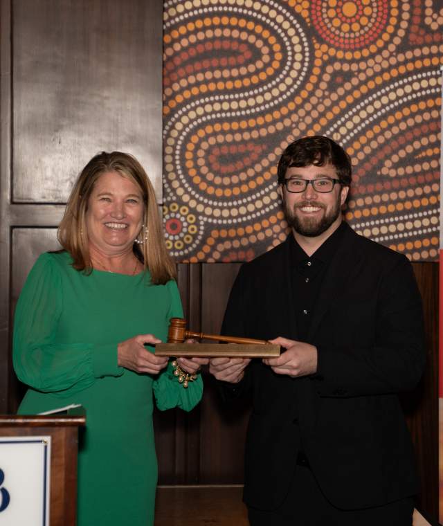 A woman in a green dress and a man in black attire smile while holding a wooden gavel together during an award or recognition ceremony. They stand beside a podium with the Destination NMB logo and a banner featuring sponsor information. The background features a decorative patterned wall.