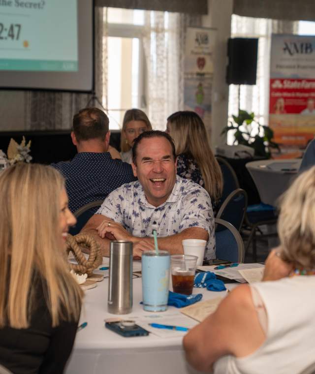 People seated around round tables engage in lively conversation during the Marketing Outlook Summit. One man at the center smiles and laughs while talking with others. A large screen with a countdown timer and banners with the NMB logo are visible in the background, indicating a timed group activity or networking session.