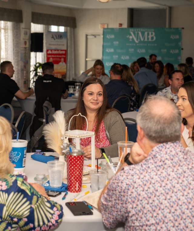 Groups of people sit around round tables engaged in discussion at a professional event or workshop. A teal “Destination NMB” backdrop is visible in the background along with projected slides and promotional banners. The atmosphere is collaborative and social, with attendees smiling and interacting across the room.