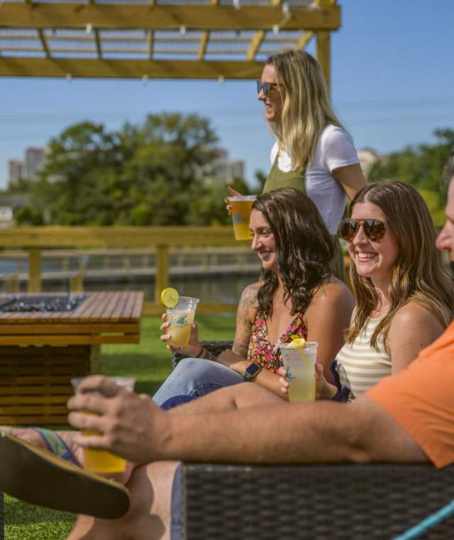 Group of friends relaxing with drinks and appetizers on a sunny outdoor patio in North Myrtle Beach, South Carolina, enjoying casual coastal dining and vibrant social atmosphere under a pergola.