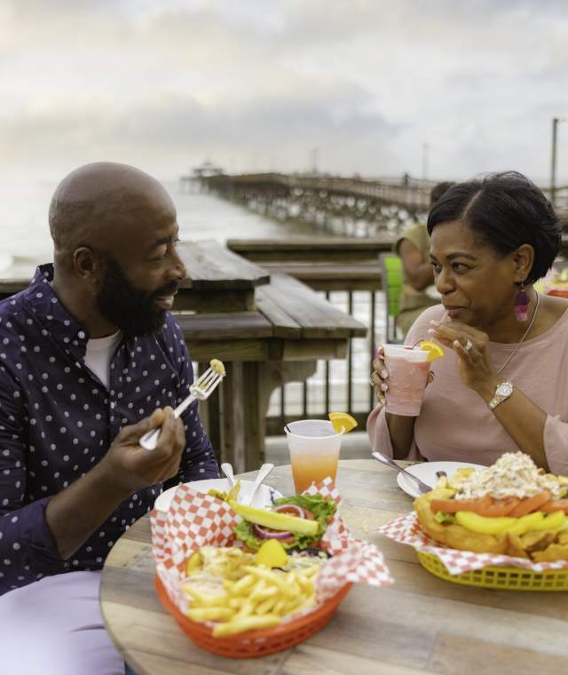 Smiling couple enjoying food and drinks at an outdoor beachside restaurant, with a pier and ocean in the background.