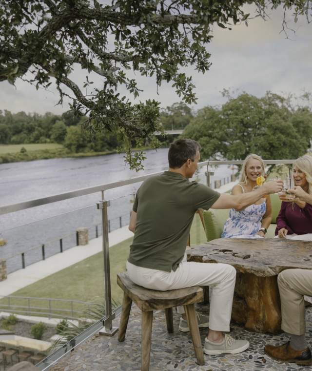 Four adults sit on a riverside terrace, smiling and clinking glasses around a wooden table. Boats pass by on the water below, and trees and buildings are in the background.