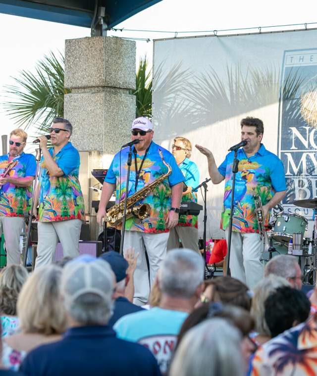 A band performs on an outdoor stage in North Myrtle Beach, wearing colorful tropical shirts and entertaining a large crowd. One musician plays a saxophone while others sing and play guitar, drums, and keyboard. The audience watches enthusiastically. A banner in the background features the North Myrtle Beach logo.