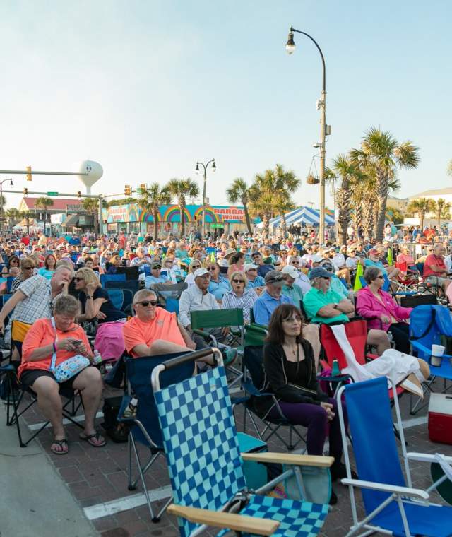 A large crowd sits in folding chairs along a street in North Myrtle Beach, enjoying an outdoor event on a sunny day. Palm trees, tents, and colorful shops line the background.