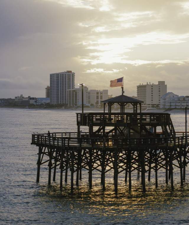 Cherry Grove Pier extends over the ocean in North Myrtle Beach. The American flag waves atop the pier’s iconic octagonal pavilion, and the sun reflects off the water below. A few people are seen fishing along the pier, while the shoreline in the background is lined with coastal homes and high-rise buildings.