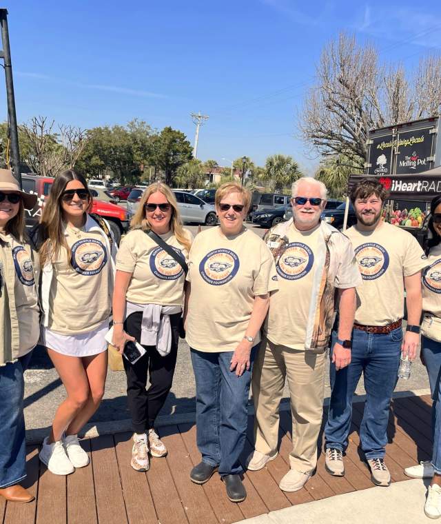 Seven people wearing matching event T-shirts stand smiling outdoors in sunny weather. They pose together in front of vendor trucks and an iHeartRadio tent at the inaugural Oyster Roast for the Coast.