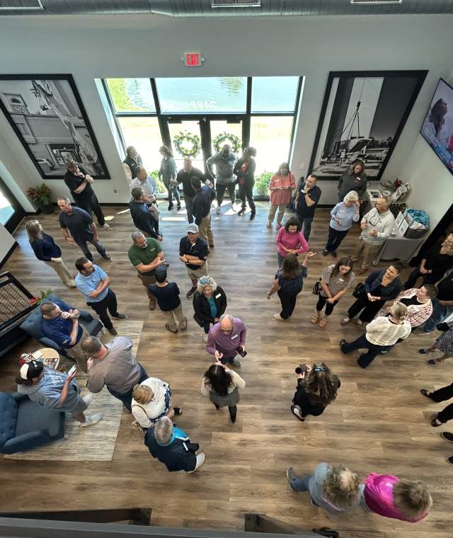 Overhead view of a group of people gathered in a spacious lobby with wood flooring and modern decor. Guests are mingling and conversing near the entrance, where sunlight streams through large glass doors. The setting appears to be a professional or community event in a welcoming indoor space