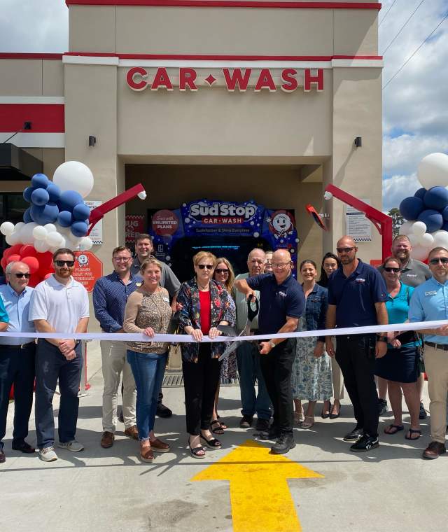 A group of chamber partners and ambassadors stand in front of a newly opened car wash for a ribbon-cutting ceremony. The building is decorated with red, white, and blue balloons, and a large ribbon is being held across the entrance. Smiling participants include business representatives and community members, celebrating under a partly cloudy sky