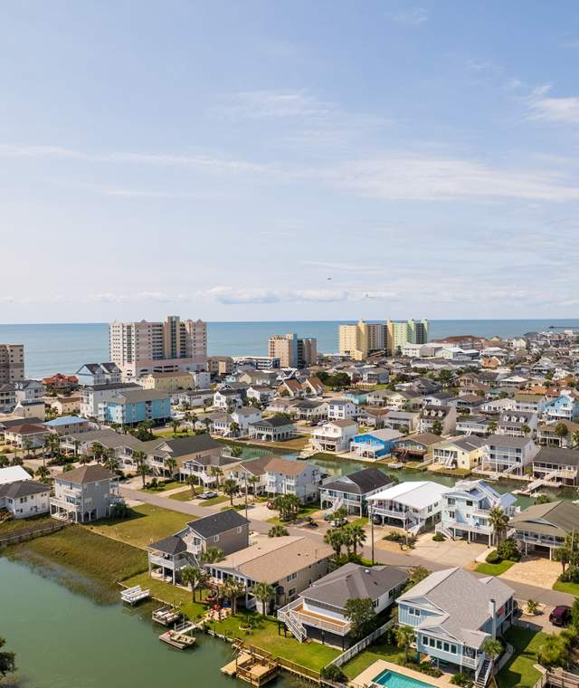 Aerial view of North Myrtle Beach featuring waterfront homes along canals in the foreground, with high-rise hotels and condos near the shoreline. The Atlantic Ocean stretches across the horizon under a partly cloudy sky.