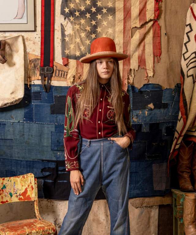 A person poses with one hand by their side and one in their pocket while wearing contemporary versions of traditional fashion clothing, standing in front of a wall adorned with many eclectic items and clothing