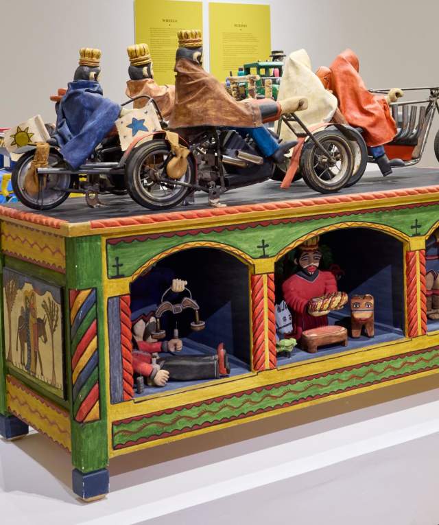 Art installation featuring vibrantly painted toy-like motorcycles on top of a colorful display case with detailed figures inside, exhibited in a gallery setting.