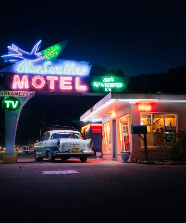 Night view of the Blue Swallow Motel with vibrant neon signs, featuring a classic car parked in front.