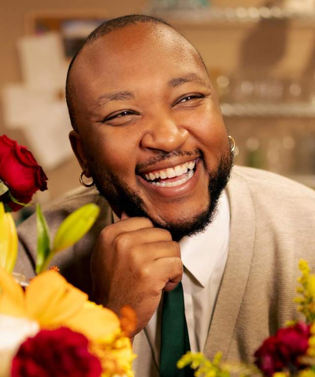 Florist in a flower shop, smiling warmly, surrounded by colorful bouquets of roses and other flowers.