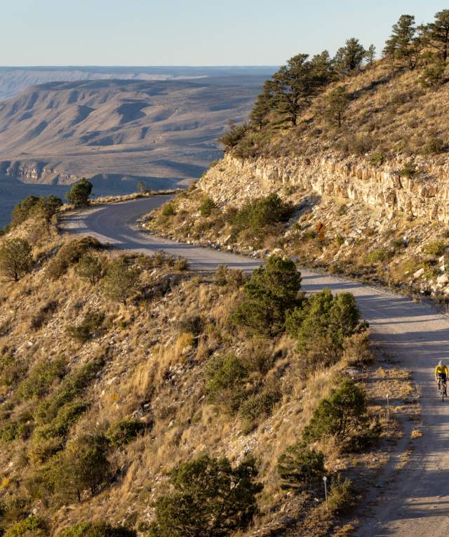 A person cycling on a winding mountain road at sunset, with expansive views of layered hills in the background.