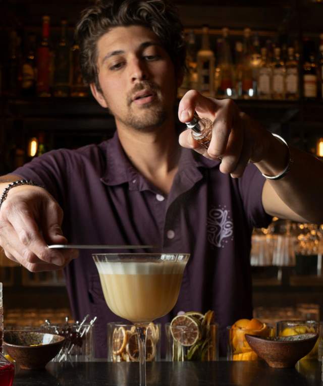A bartender garnishing a cocktail at a bar, surrounded by an array of bottles and ingredients.