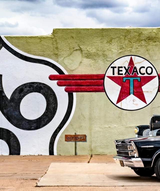 A vintage car is parked in front of a wall painted with a large "Route 66" emblem and a Texaco logo. Two people stand near the wall. The scene conveys a classic, nostalgic feel.