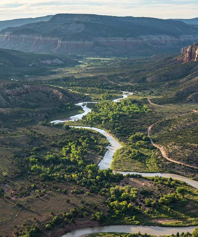 a river runs through a lush green landscape between rolling mountains and cliffs, trails curving around to either side.