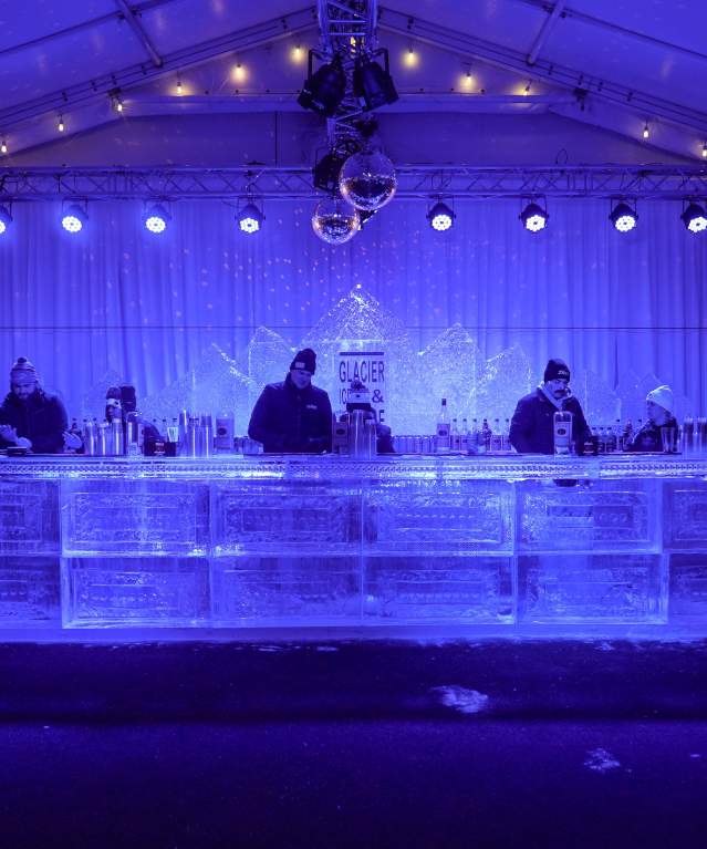 Several bartenders serving drinks at a bar made of ice under deep blue lights.