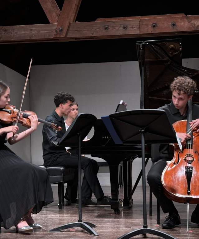 Four Luzerne Music Center students playing violin, cello and piano on stage indoors with white wall in back
