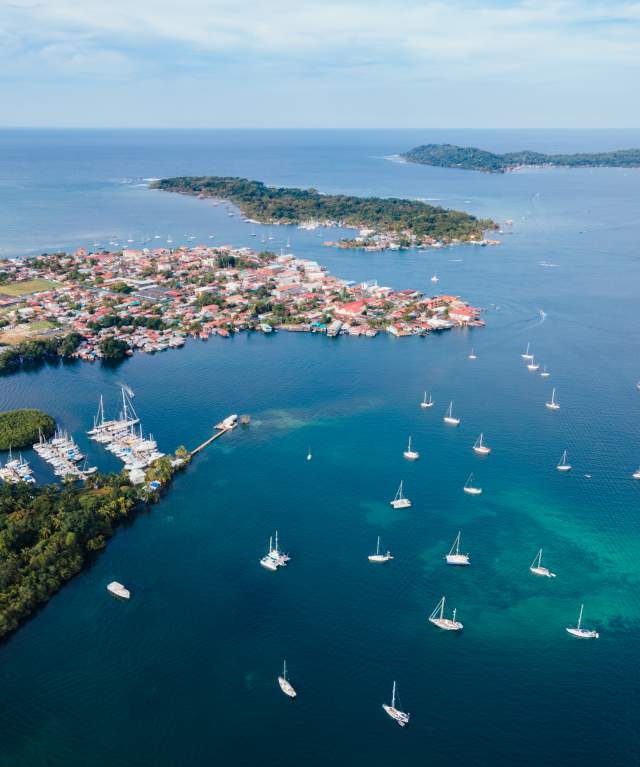 An aerial view shows a vibrant coastal town with colorful, red-roofed buildings situated among lush green islands and a tranquil bay filled with sailboats. A marina and a small airport are also visible, suggesting a popular sailing destination.