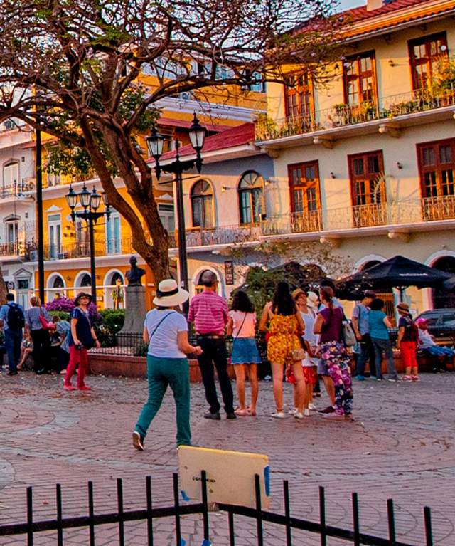 The image shows a vibrant street scene in a historic city. The street is lined with colorful buildings, some with balconies and ornate details. There are people walking around, enjoying the evening. In the background, there is a church with a blue facade and a bell tower.