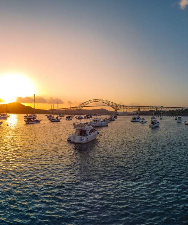 Beautiful sunset over a harbor. The sun is setting behind a bridge of the Americas in the distance, casting a golden light on the water. There are many boats anchored in the harbor, and people can be seen walking along the docks.