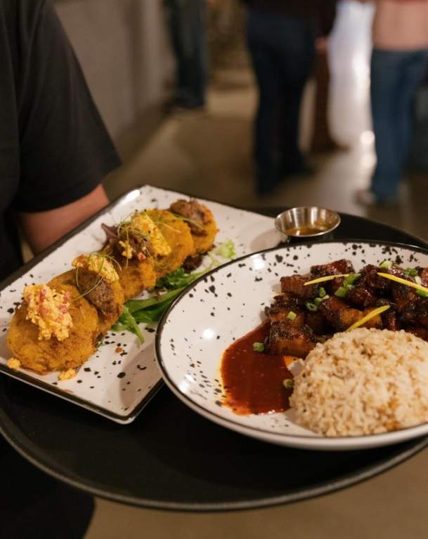 A server holds a black tray featuring two plates: one with golden fritters topped with herbs, and another with glazed meat, rice, and sauce.