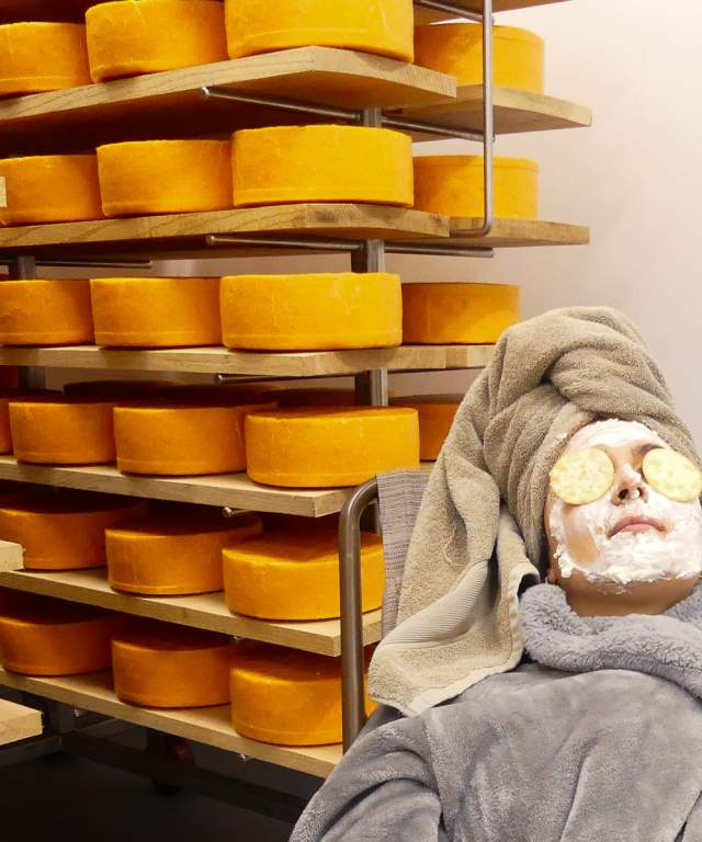 A person reclined in front of wheels of cheese in an aging room with a crowdie cheese face mask and crackers on their eyes, wearing a robe and towel on their head