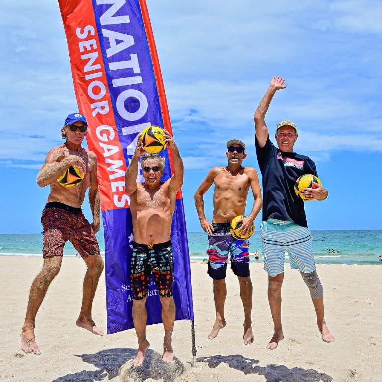 Four men jumping in the air on Fort Lauderdale Beach holding volleyballs in front of the National Senior Games flag