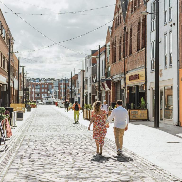 Humber Street on a sunny day facing out to the marina. The brightly coloured shop fronts lead into the distance where you can just see boats bobbing on the water. Lights are strung across the road from roof to roof. A couple strolls away from the camera, the lady wears a summer dress and the man is in wearing light chinos and a white shirt.