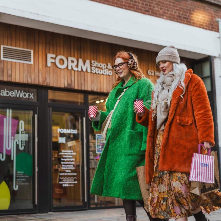 Two women walk down Humber Street's cobbles. They stroll past FORM Shop's wooden exterior with a colourful display in the window. They are bundled up in hats and scarves and are carrying coffees and shopping bags.