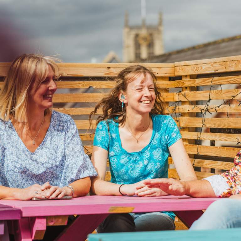 A man and woman  wearing a hearing aid relaxes group of friends at a colourful outdoor seating area on Humber Street in Hull. The scene showcases the vibrant, accessible social spaces of the Humber Street area