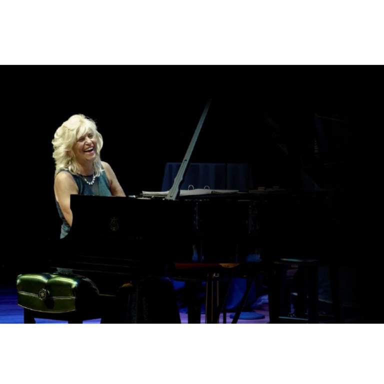 Jodie DeSalvo a smiling blonde haired woman, sits at a piano while laughing.