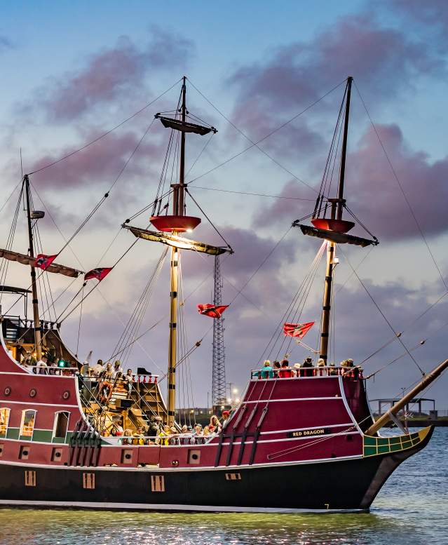 Large red pirate ship in the ocean with lights on in facing the deck. The sun has almost set and gives the sky and clouds a purple tint.