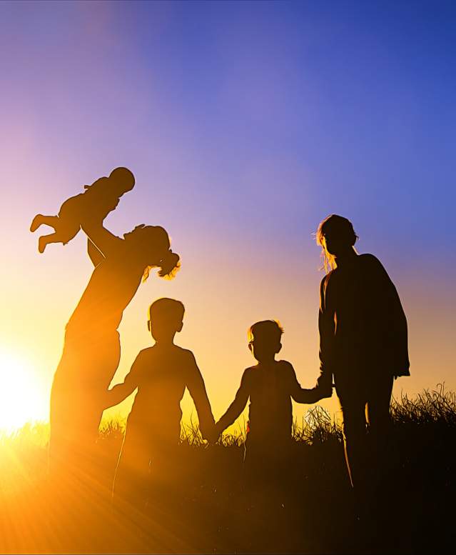A family is silhouetted against the sun. A mother holds a baby in the air, while three other children hold hands next to them